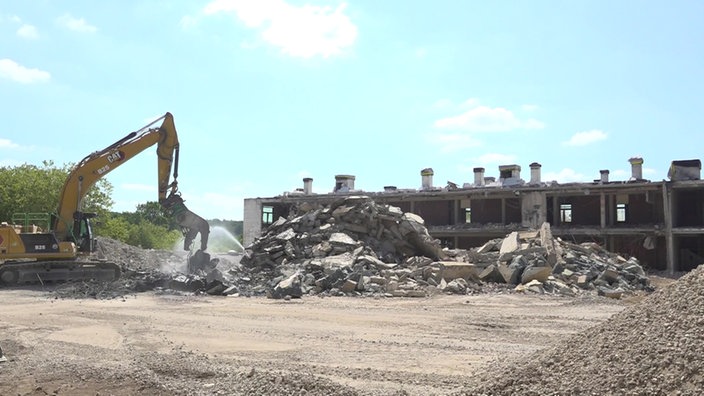 Ein Bagger vor einem Haufen Schutt, im Hintergrund noch die Ruine eines Gebäudes