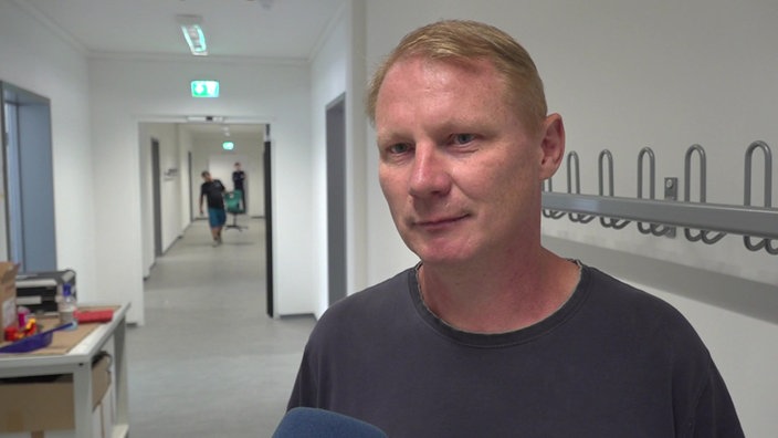 Ein Mann in einem dunklen T-Shirt steht in einem langen Gang mit Türen und Kleiderhaken