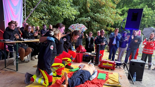 Menschen stehen auf einer Bühne, eine Übungspuppe wird reanimiert