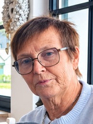 Eine Frau mit Brille und dunklem Kurzhaarschnitt sitzt in einem Gastraum. Im Hintergrund sind Sprossenfenster zu sehen.