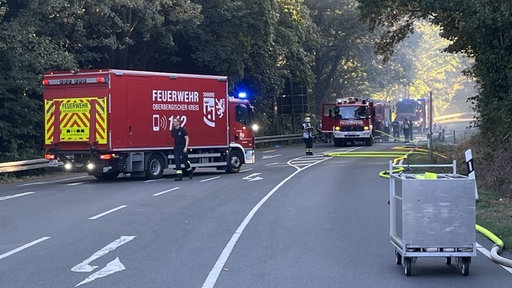 Einsatzkräfte, Fahrzeuge und Wasserschläuche auf einer Straße in einem Waldstück bei Lindlar