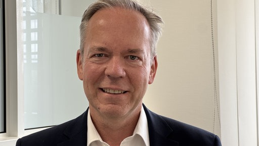 Ein Mann mittleren Alters lächelt in die Kamera. Er hat einen Kurzhaarschnitt und trägt ein weißes Hemd mit einem dunklen Jackett.