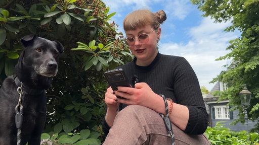 Angie vom CSD Oberberg sitzt draußen zwischen Bäumen und schaut auf das Smartphone in ihrer Hand.