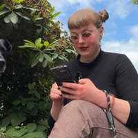 Angie vom CSD Oberberg sitzt draußen zwischen Bäumen und schaut auf das Smartphone in ihrer Hand.