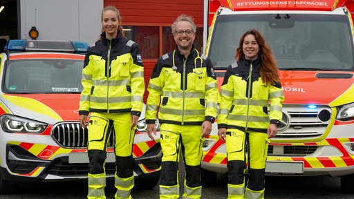Drei Menschen in gelber Einsatzkleidung vor zwei Fahrzeugen des Rettungsdienstes.