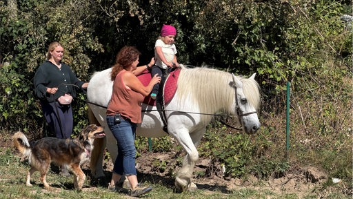 Ein kleines Mädchen reitet auf einem Schimmel und wird von zwei Frauen geführt