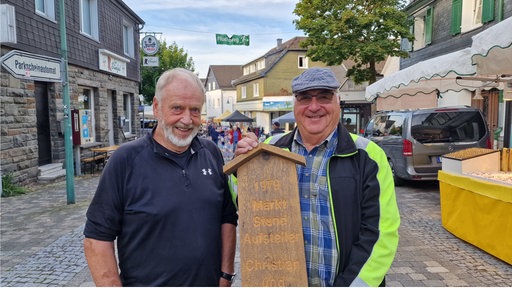 Zwei Männer stehen zwischen Marktständen und lächeln.