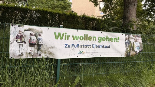 An einem Zaun hängt ein Banner mit der Aufschrift "Wir wollen gehen! Zu Fuß statt Elterntaxi. Kreis Mettmann".