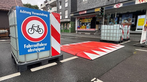 Auf einer Straße steht ein Wassercontainer mit einem Schild: Achtung Veranstaltung. Daneben auf dem Boden Straßensperren aus Metall.