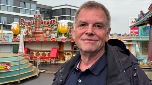 Ein Mann steht auf einer Kirmes und lächelt in die Kamera.