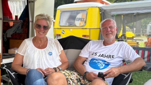 Ein weißhaariger Mann mit Brille und eine Frau mit blinden kurzen Haaren und Sonnenbrille sitzen an ihrem Wohnwagen und lächeln.