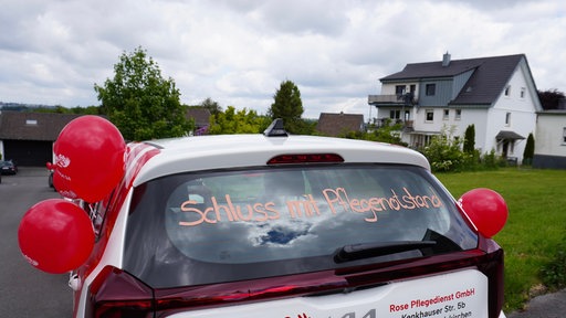 Weißes Auto mit Luftballons, auf dem Heckfenster steht "Schluss mit Pflegenotstand"