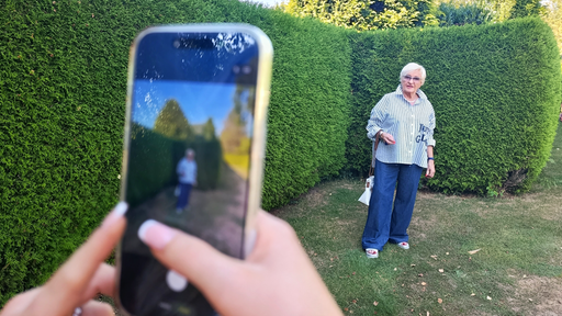 Eine ältere Dame steht in einem Garten vor einer Hecke und wird mit einem Smartphone fotografiert.