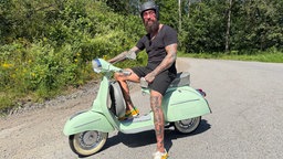 Ein Mann mit Bart sitzt auf einer grünen Vespa
