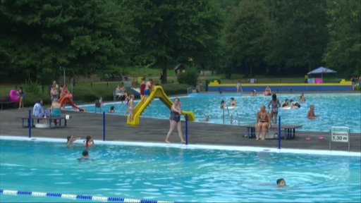 Menschen baden und entspannen in einem Freibad mit mehreren Becken und Wasserrutschen.