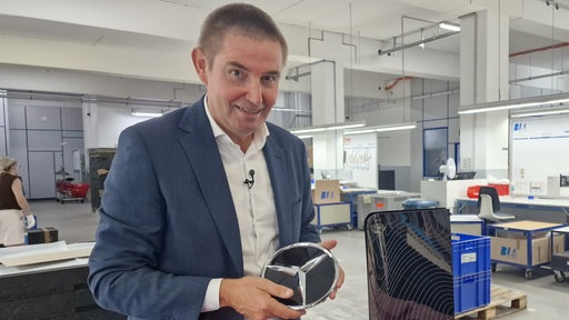 Ein Mann hält einen Mercedes-Stern in der Hand, daneben steht eine verchromte Schutzhülle für die Playstation.