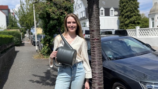 Laura Hertel steht mit einer Gießkanne vor dem Baum