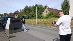 Eine junge Frau neben einem großen Blitzer. Ein Mann fotografiert sie.