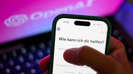 Ein Mensch hält ein Handy in der Hand, auf dem Chat-GPT geöffnet ist. Die App fragt "Wie kann ich dir helfen?" Im Hintergrund ein aufgeklappter Laptop, der das "Open AI"-Logo zeigt