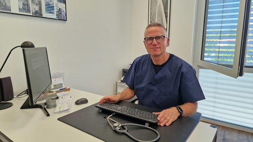 Ein Arzt im blauen Kittel sitzt an seinem Schreibtisch und schaut in die Kamera, vor ihm eine Tastatur und ein Stethoskop