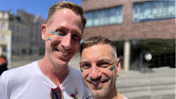 Zwei Männer stehen Arm in Arm und lächeln. Sie haben Regenbogenflaggen auf die Wangen gemalt.