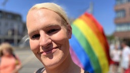 Blonde Frau lächelt in die Kamera, Regenbogenflagge im Hintergrund
