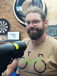 Ein Mann mit dunklen Haaren und Vollbart steht in einer Fahrradwerkstatt. Er trägt ein T-Shirt mit einem Fahrradmotiv. Im Hintergrund hängt verschiedenes Fahrradzubehör an einer Holzwand.