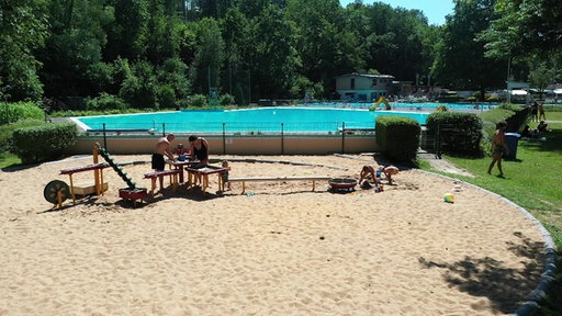 Sandspielbereich mit spielenden Kindern vor einem Freibad mit großem Schwimmbecken und Bäumen im Hintergrund