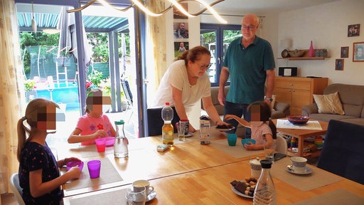 Blonde Frau und ihr Mann vor dem Esstisch, an dem drei Kinder sitzen, den sie gerade Getränke einschüttet