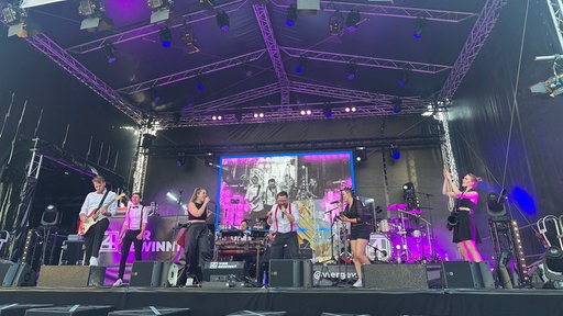 Frauen und Männer stehen gemeinsam auf einer Bühne, singen und musizieren.