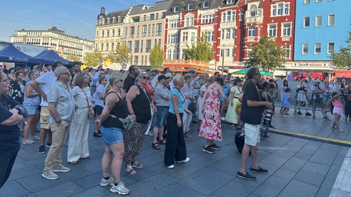 Menschen mit Sommerkleidung und Sonnenbrillen stehen als Publikum auf einem Festgelände