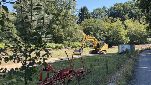 Ein Bagger auf Wiese, im Hintergrund viele Bäume