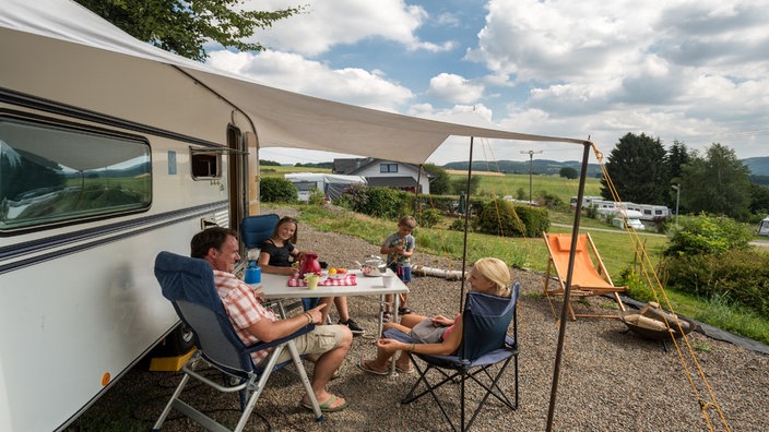 Eine Familie sitzt auf einem Campingplatz unter einem Sonnensegel und unterhält sich.