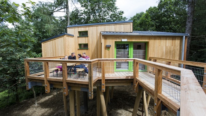 Eine Familie sitzt auf einem Baumhaus an einem Holztisch und unterhält sich.