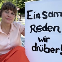 Eine Frau hält ein Schild auf dem steht: "Einsam? Reden wir drüber!"
