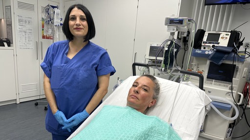 Eine Gesundheitspflegerin im blauen Kasack steht an einem Krankenbett in dem eine Patientin liegt und mit einem grünen Tuch bedeckt ist.