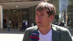 Ein Mann, im Interview mit dem WDR, steht vor einem Gebäude mit Schaufenstern