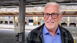 Ein Mann mit Brille und kurzen grauen Haaren steht auf einem Bahnsteig.