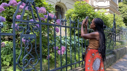 Eine Frau hat das Handy in der Hand und filmt damit ein Haus und Blumen