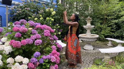 Eine Frau hat das Handy in der Hand und filmt damit ein Haus und Blumen