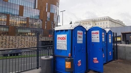 Blaue mobile Toilettenhäuser an einer Straße, im Hintergrund ein großes Gebäude mit einer Glas- und Kupferfassade.