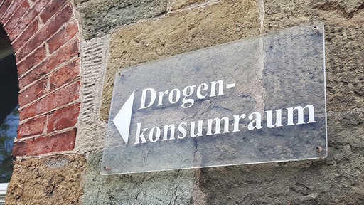 Ein Hinweisschild mit der Aufschrift „Drogenkonsumraum“ ist an der Fassade eines alten Natursteingebäudes mit einem Rundbogen aus rotem Backstein angebracht.