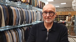 Ein Mann mit Glatze und Brille steht in einem Bekleidungsgeschäft für Herrenmode