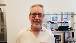 Ein Mann mit Brille und grauen Haaren, bekleidet mit einem beigen Poloshirt, steht in einem Optikergeschäft.