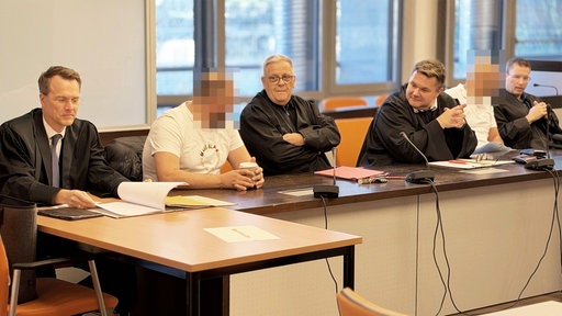 Zwei angeklagte Männer in weißen Hemden sitzen gemeinsam mit vier männlichen Verteidigern an einem Tisch im Gerichtssaal.