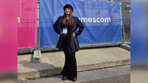 Eine Frau in schwarzer Kleidung und mit einem Hut steht vor einem Gamescom-Banner.