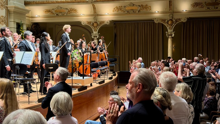 Patrick Hahn steht mit dem Orchester auf der Bühne, das Publikum klatscht