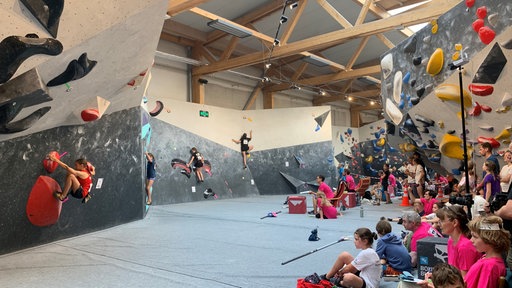 Mehrere Kinder auf einer Kletterwand mit Zuschauern am Bildrand.