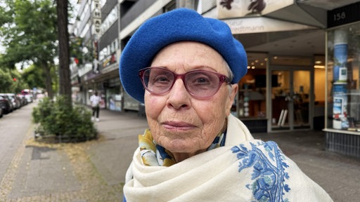 Eine Frau mit Brille und blauer Baskenmütze steht an einer Einkaufsstrasse.