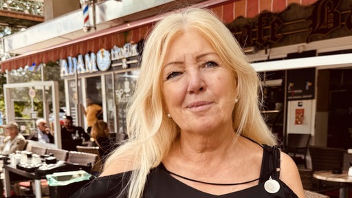 Eine Frau mit langen blonden Haaren, bekleidet mit einem schwarzen Oberteil, steht vor einem Eiscafé.
